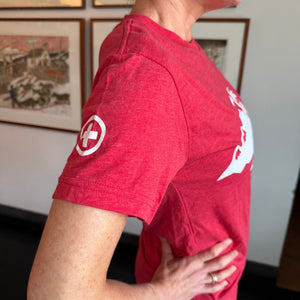 Person wearing a red t-shirt with a logo, standing indoors with framed pictures on the wall.