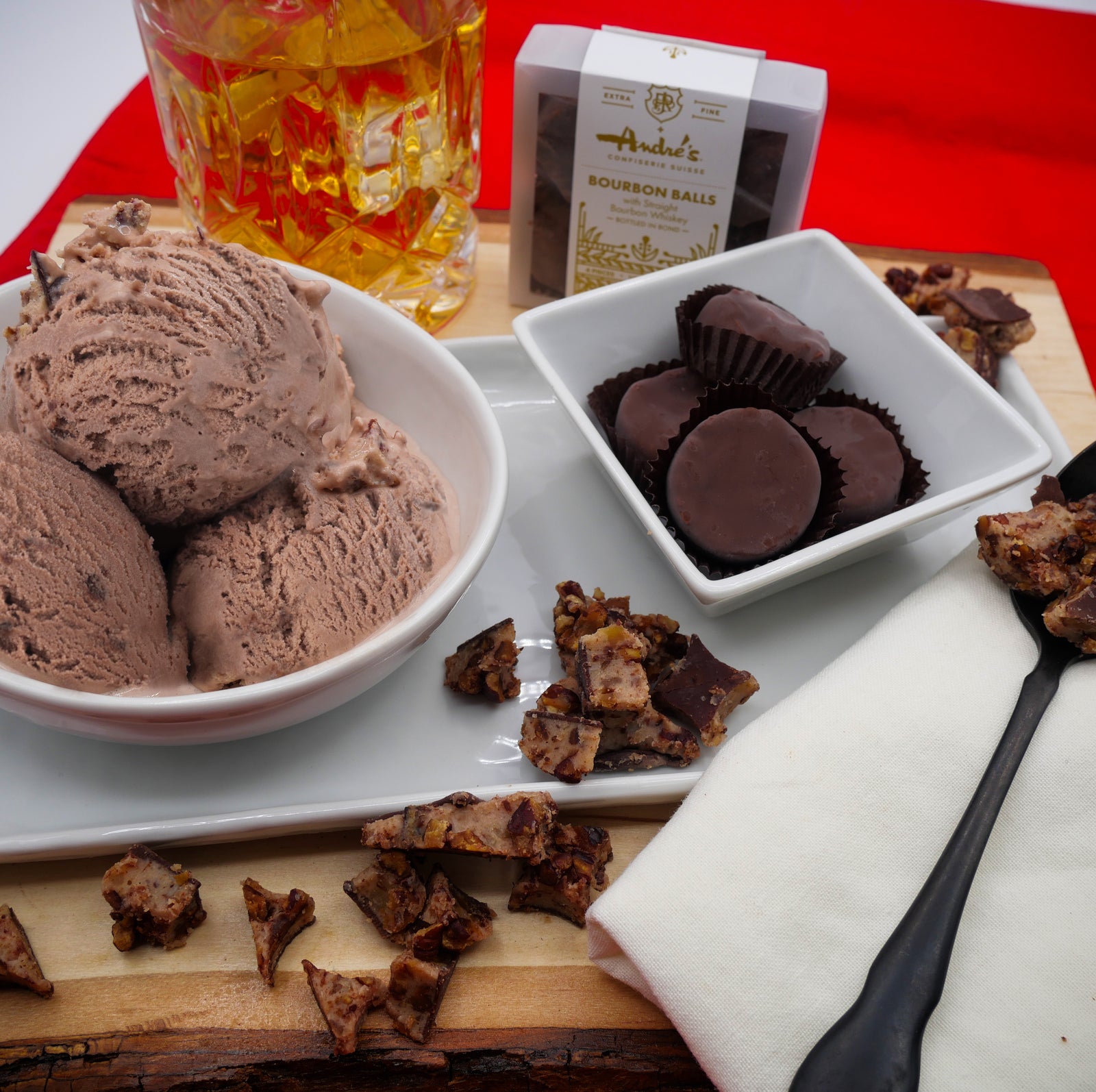 glass of bourbon, box of bourbon balls, bowl with scoops of Clementine's Bourbon Balls Ice Cream with loose and cut up Bourbon Balls scattered around