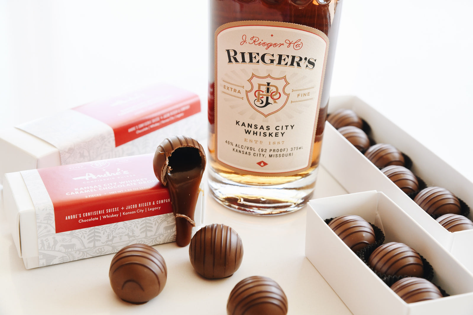 Bottle of Rieger's Kansas City Whiskey with boxed and loose whiskey caramel candies around. One is broken open and oozing caramel onto the table