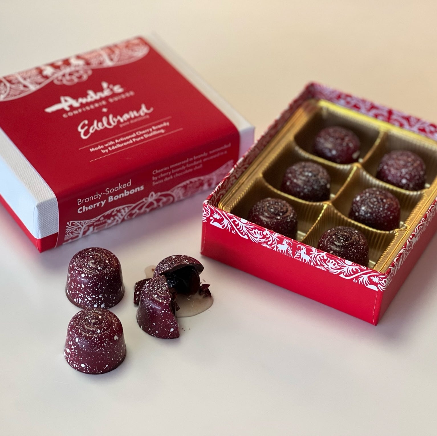 red open box base with 6 maroon and red speckled candies inside, next to a closed box with white lid and red label and loose candies, one cut open with brandy pouring from it onto the counter.