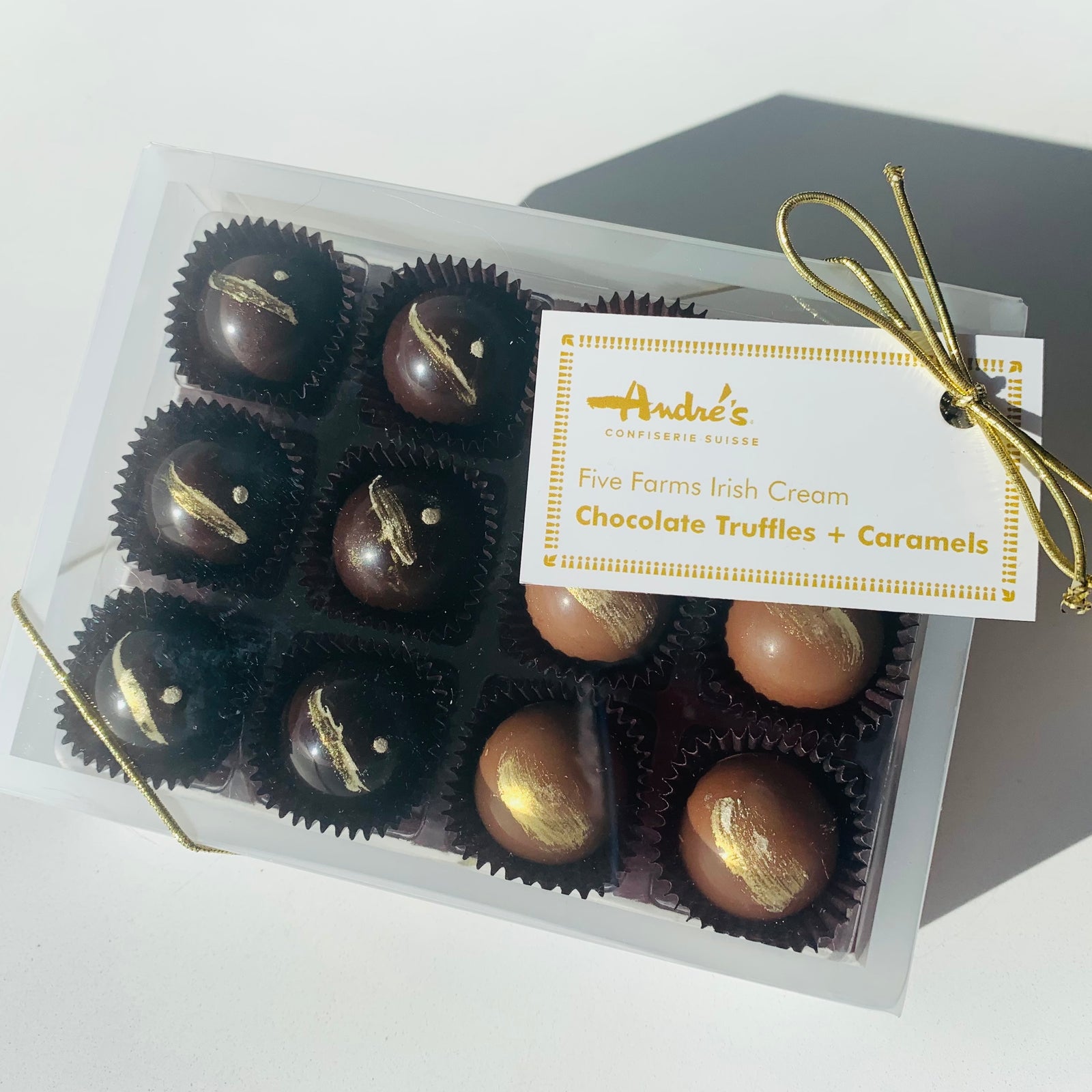 clear box with 6 round dark chocolate truffles and 6 round milk chocolate caramels, all with gold stripes, with a gold ribbon and gold and white product tag on a white table