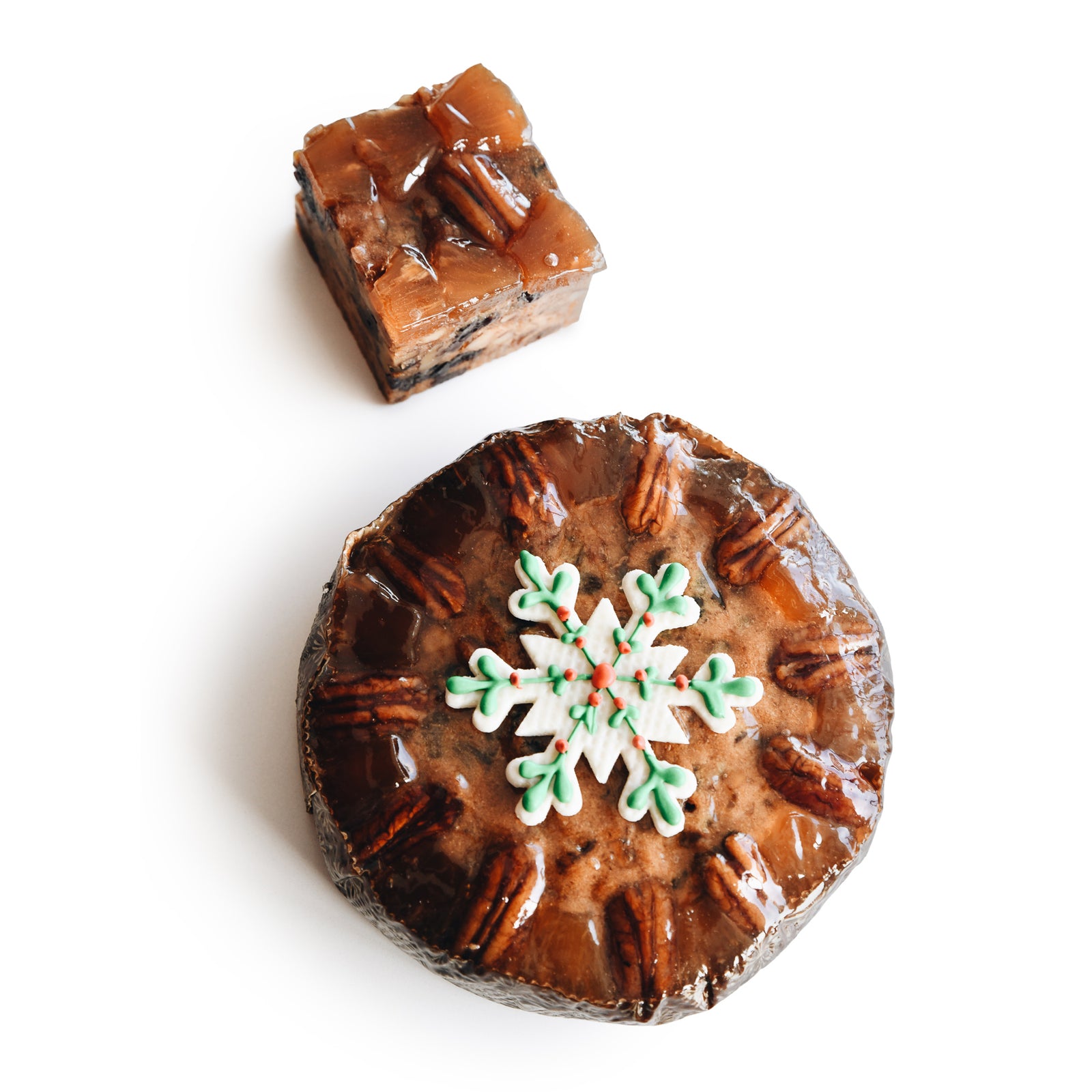 A round fruitcake with visible pecans around the edge on a gold scalloped base and round white candy disk that has candy holly and poinsettia next to a square fruitcake in a rectangular box.