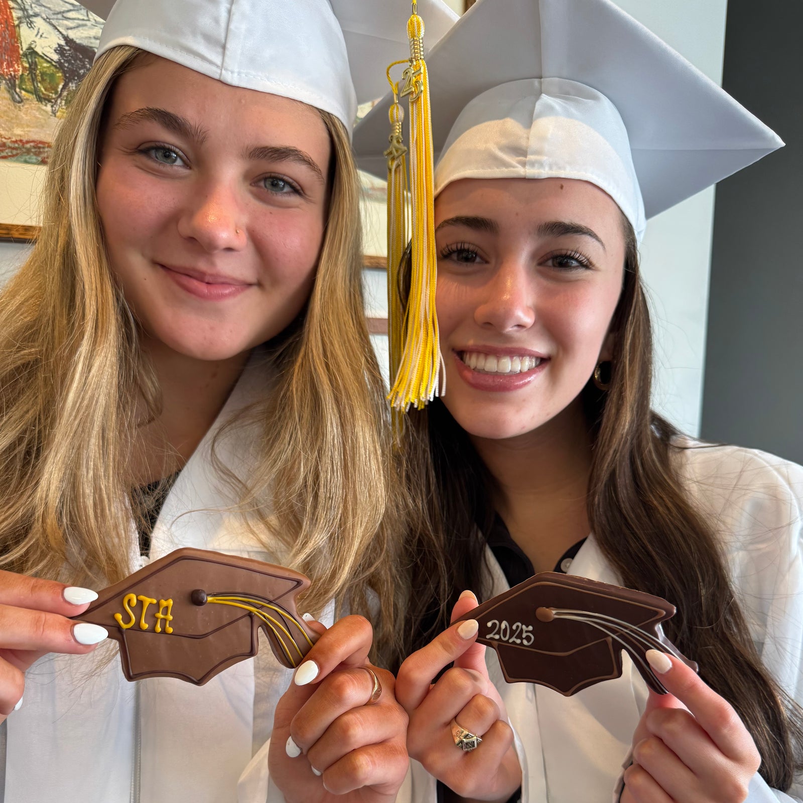 Several chocolate coated cookies shaped like graduation mortarboard caps, each with either a name, school initials, or 2025 written in chocolate.