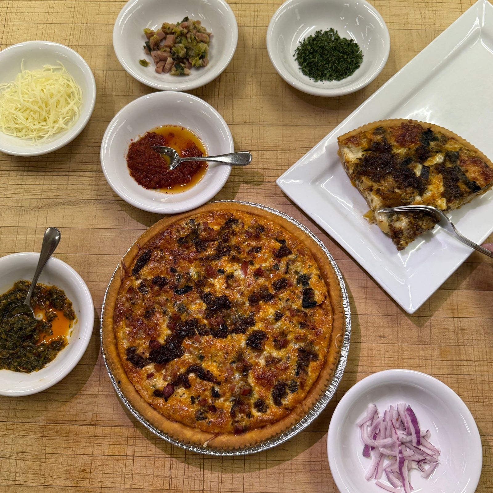 Quiche on a wooden table with ingredients in small bowls beside.