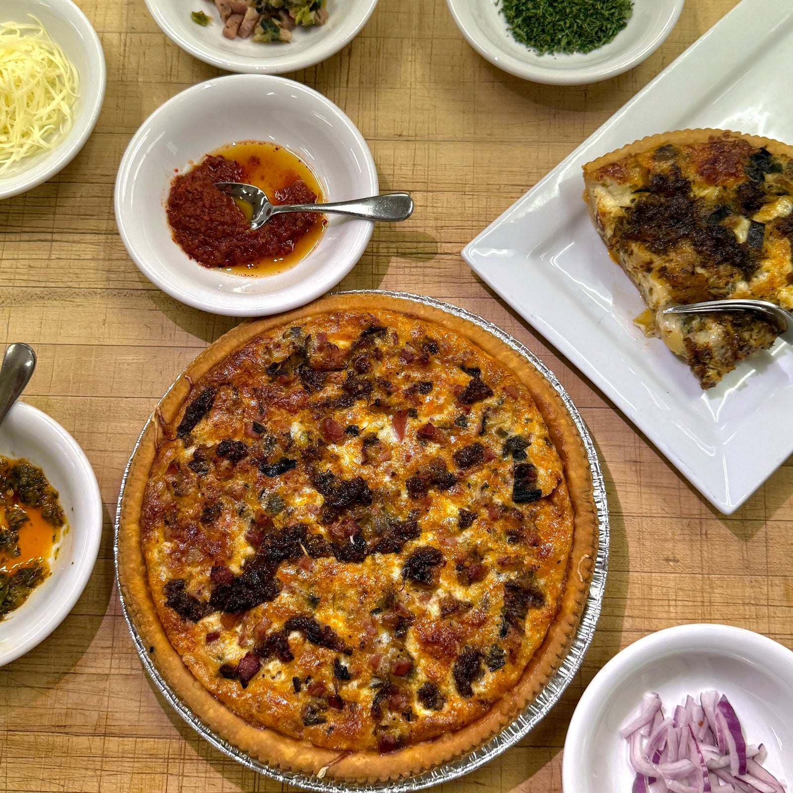 Quiche on a wooden table with ingredients in small bowls beside.
