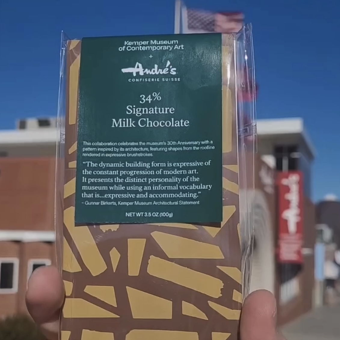 close up of Kemper Museum bar with gold abstract print in clear bag and green label that is being held up in front of the Andre's store.