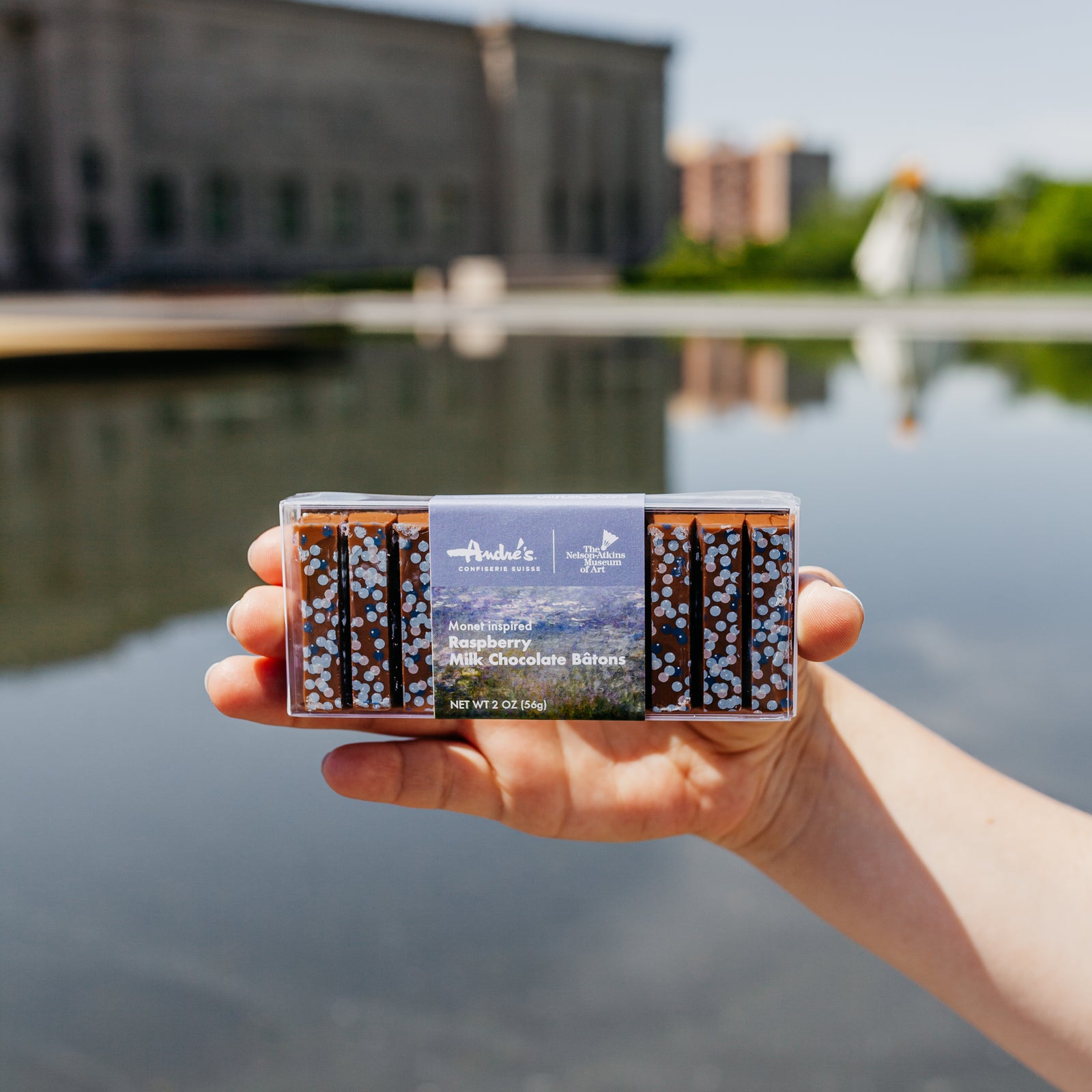 Box of the Monet Inspired Batons that feature multi-hued blue dots on the chocolate and an image of Monet's Water Lilies on the wrapper  being held up in front of the Nelson Museum.