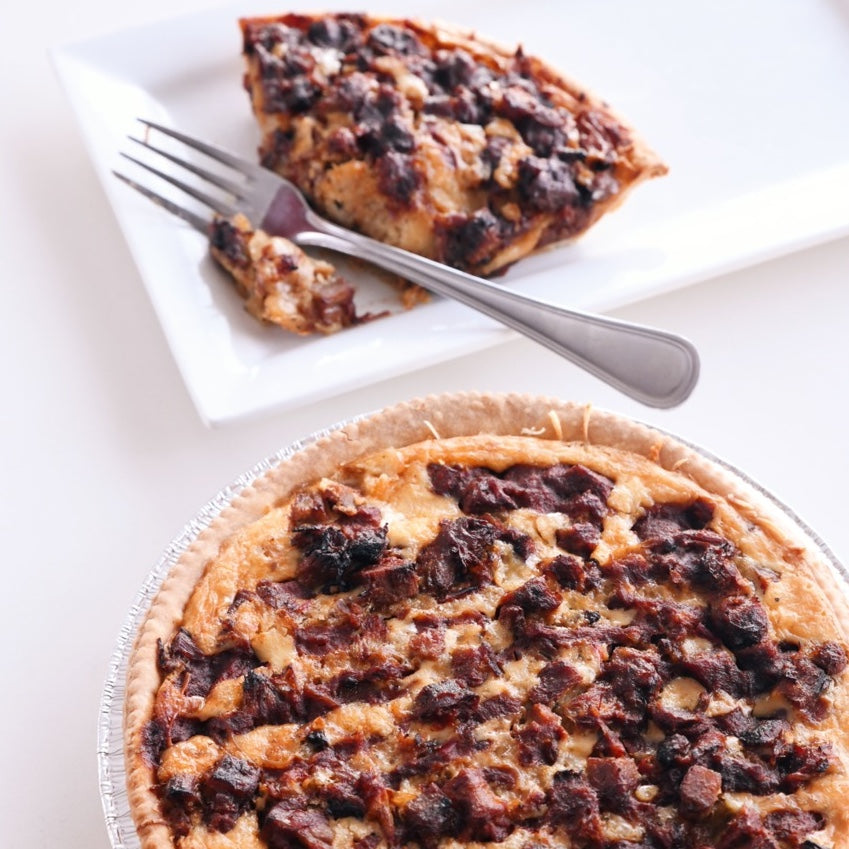 Overhead view of round quiche in a white box with golden crust and cheese with lots of brisket next to a closed box and 2 bottles of Q39 Classic Sauce.