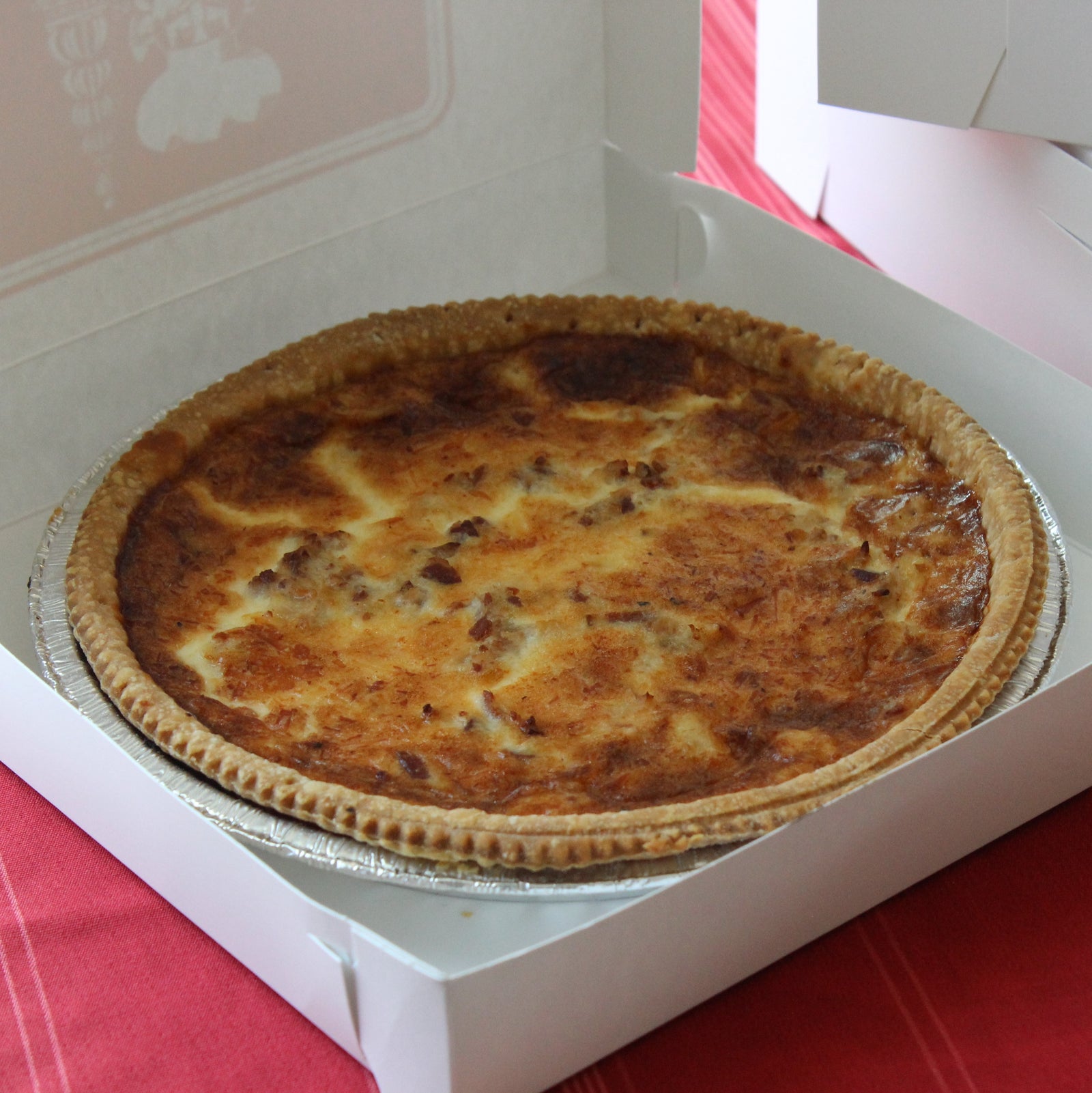 Open white quiche box holding pie pan with quiche lorraine. Golden crust and browned cheesey top with pieces of bacon visible.
