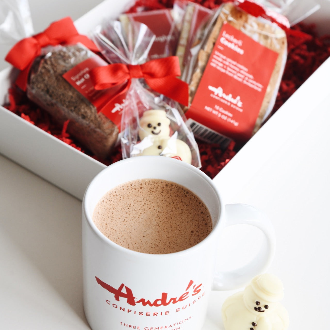 Andre's logo mug filled with hot chocolate in front of a box of assored treats in the Winter Holiday Gift Box
