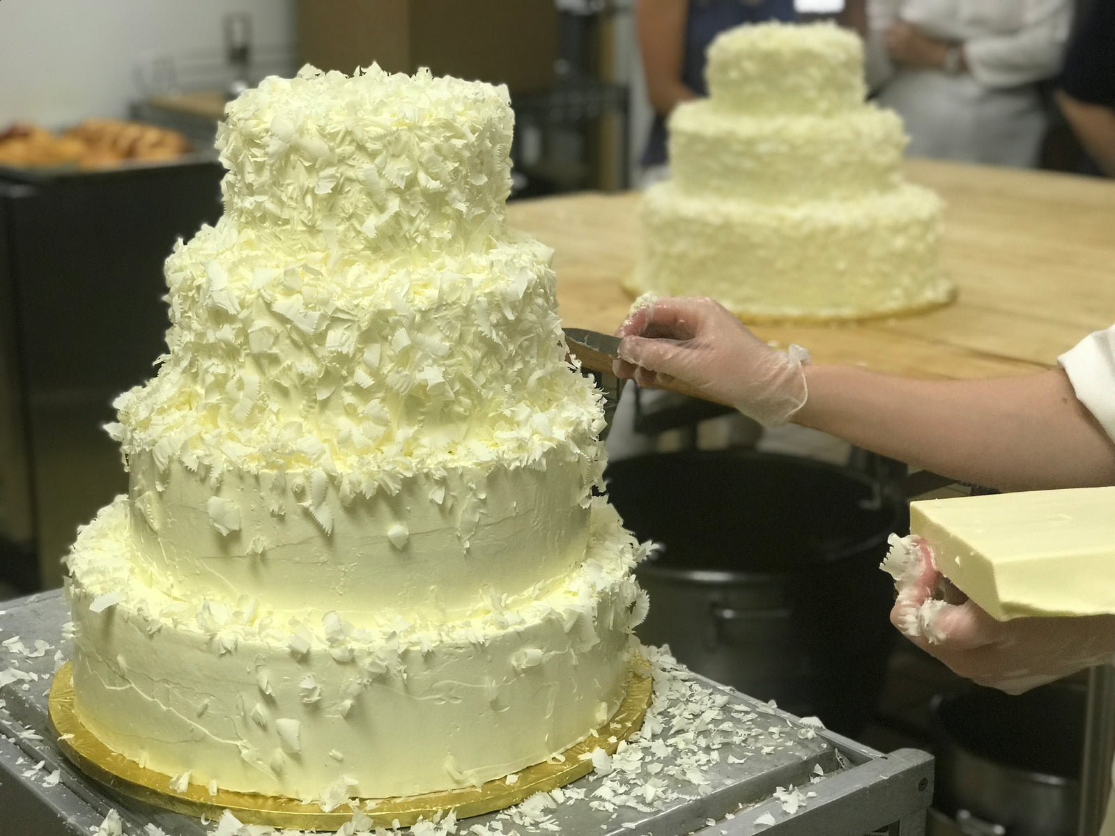 Four tiered wedding cake being covered in white chocolate shavings in front of a finished 3 tier white chocolate shavings covered cake.
