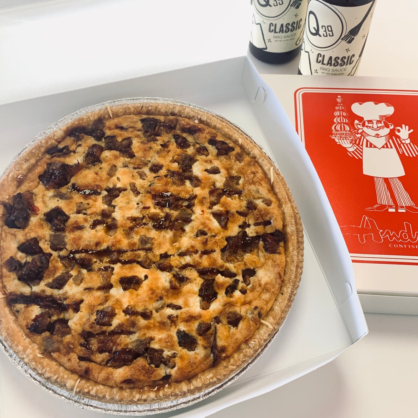 Overhead view of round quiche in a white box with golden crust and cheese with lots of brisket next to a closed box and 2 bottles of Q39 Classic Sauce.