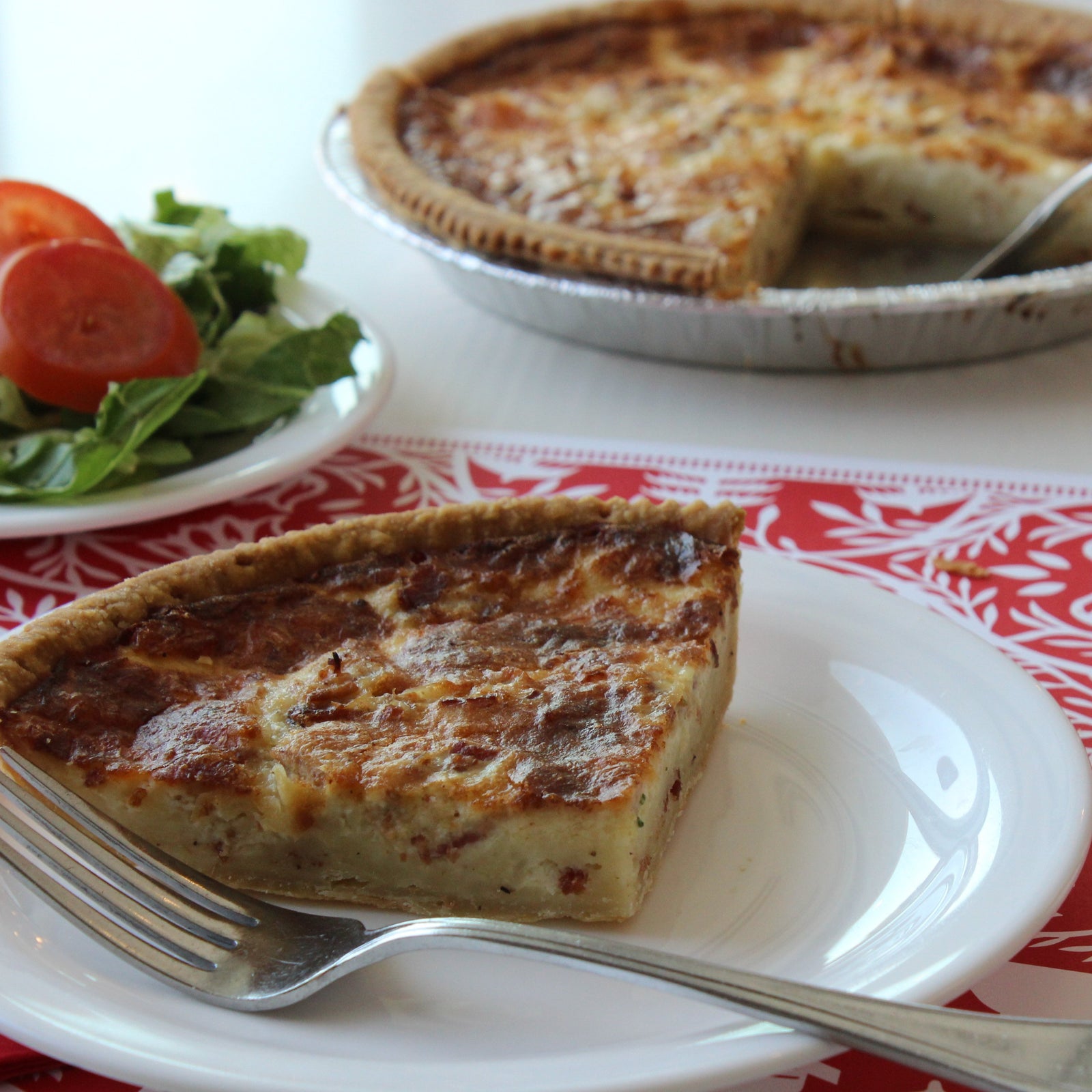 Open white quiche box holding pie pan with quiche lorraine. Golden crust and browned cheesey top with pieces of bacon visible.