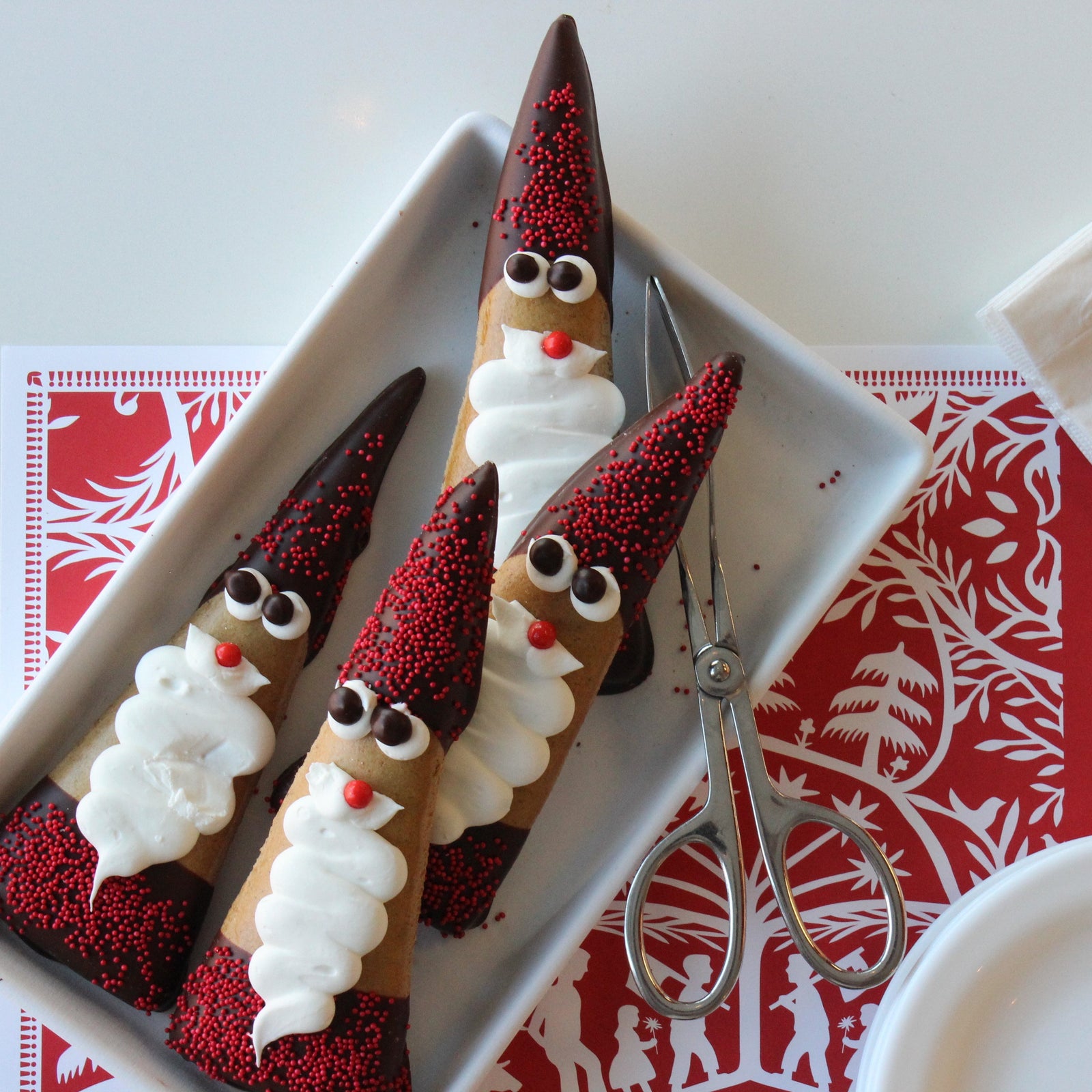 Triangular gingerbread cookie that has been dipped in dark chocolate on either end with red sprinkles to resemble cap and coat, with white icing to resemble eyes and a beard, in a bag with a red bow.