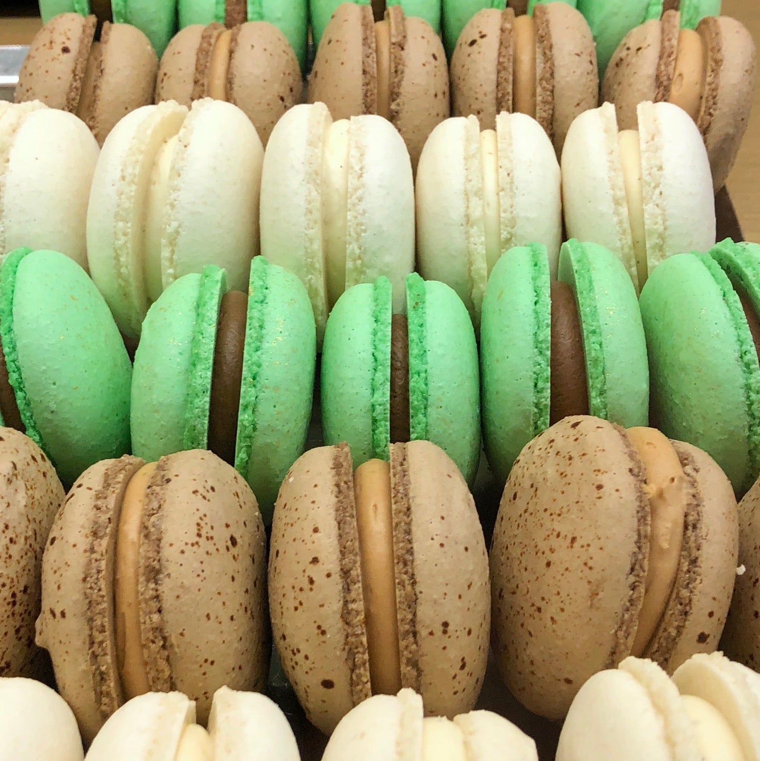 Rows of luxemburgerli cookies: white with vanilla cream center, green with chocolate cream center, speckled tan with caramel cream center.